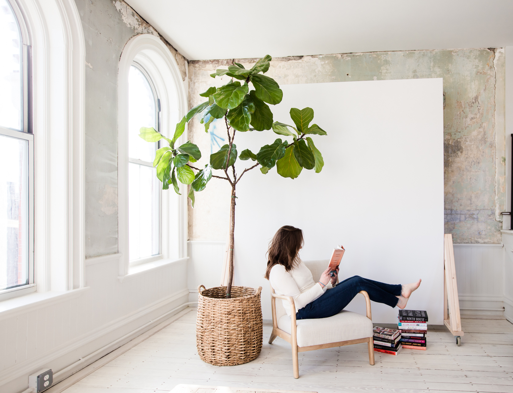 March & April Reading + Wine Pairing List | April Reading List by popular New England lifestyle blogger, Shannon Shipman: image of a woman reading a book in a white arm chair next to a fiddle leaf fig tree and pile of books.
