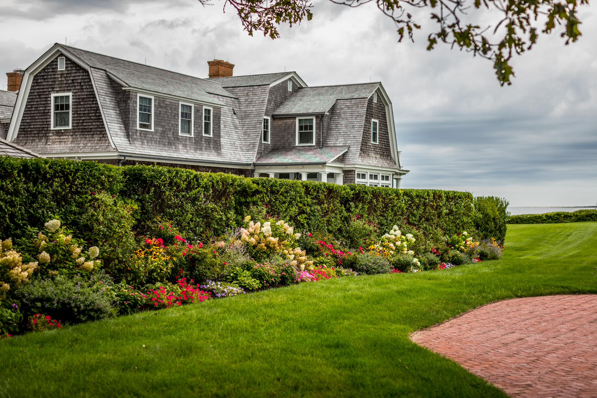 Cottage Garden Ideas by popular New England life and style blogger, Shannon Shipman: image of house with shingle siding, a hedge bush, and various flowers.