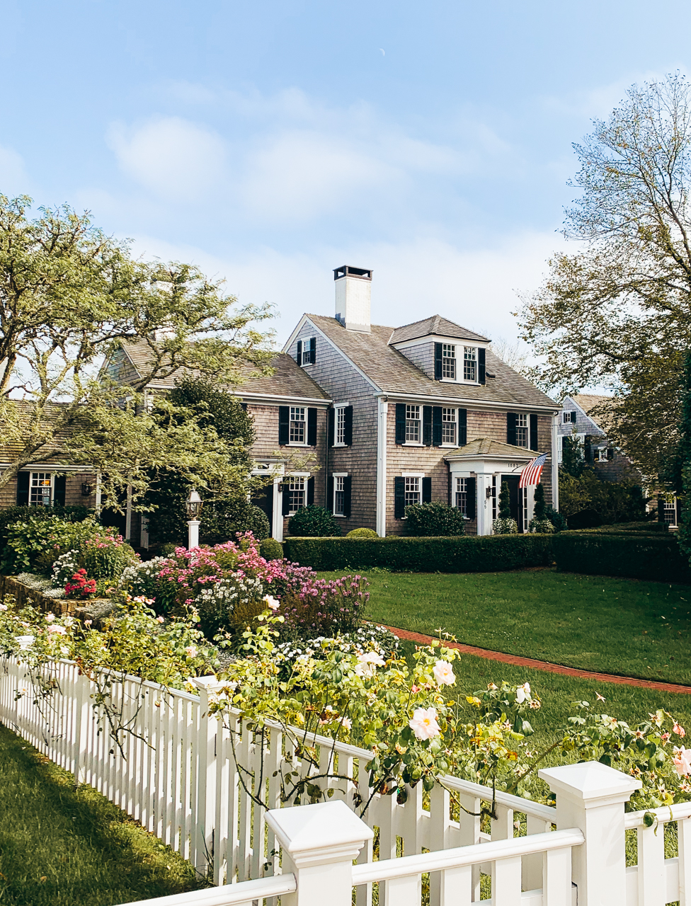 Cottage Garden Ideas by popular New England life and style blogger, Shannon Shipman: image of a colonial house with grey shingle siding and a white picket fence lined with various flowers. 